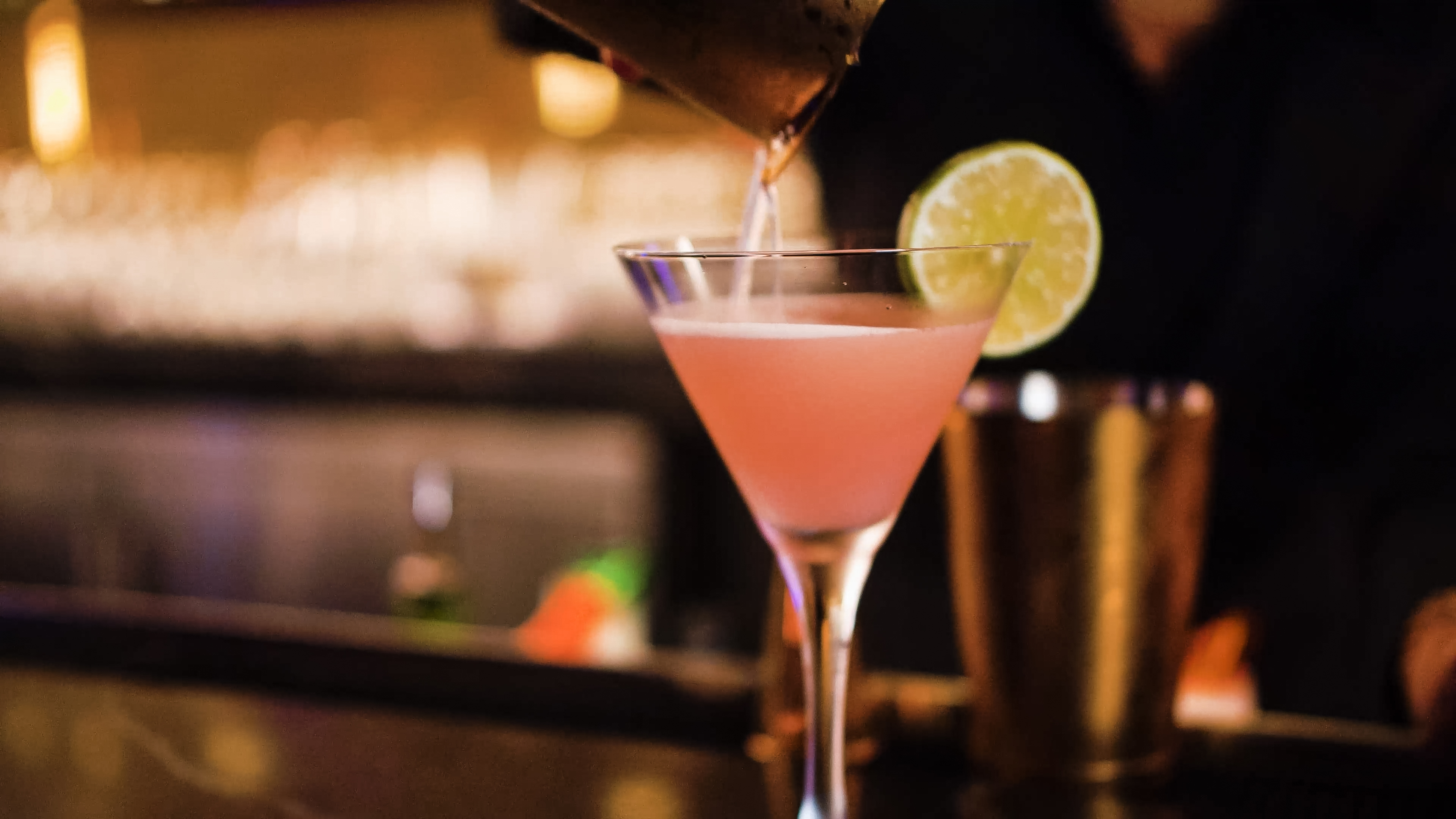 Bartender pouring a pink craft cocktail into a martini glass garnished with a fresh lime