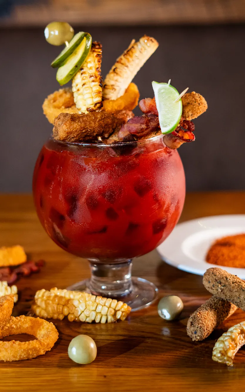 A large goblet-style cocktail overflowing with a vibrant red drink, creatively garnished with savory skewers of grilled corn, fried onion rings, mozzarella sticks, bacon, olives, and lime wedges, presented on a wooden table with scattered garnishes.