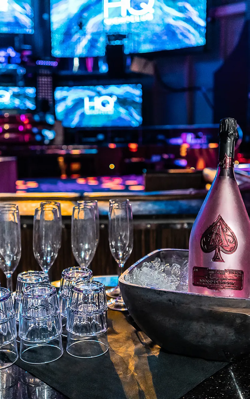A VIP table at HQ2 nightclub with a bottle of champagne on ice and glasses next to it, a lit up bar behind it