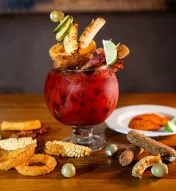 A large goblet-style cocktail overflowing with a vibrant red drink, creatively garnished with savory skewers of grilled corn, fried onion rings, mozzarella sticks, bacon, olives, and lime wedges, presented on a wooden table with scattered garnishes.