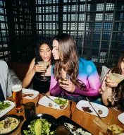 A group of young adults sipping drinks and eating at an upscale restaurant table looking happy