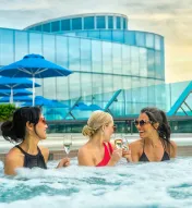 Three women sit in a rooftop jacuzzi smiling while toasting champagne