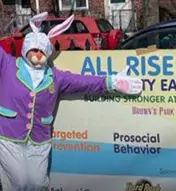 Someone dressed as an Easter bunny stands in front of a banner for an All Rise Event