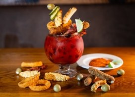 A large goblet-style cocktail overflowing with a vibrant red drink, creatively garnished with savory skewers of grilled corn, fried onion rings, mozzarella sticks, bacon, olives, and lime wedges, presented on a wooden table with scattered garnishes.