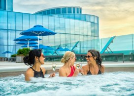 Three women sit in a rooftop jacuzzi smiling while toasting champagne